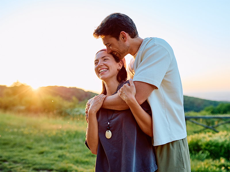 Pareja joven sonríe abrazada en un campo verde al atardecer.