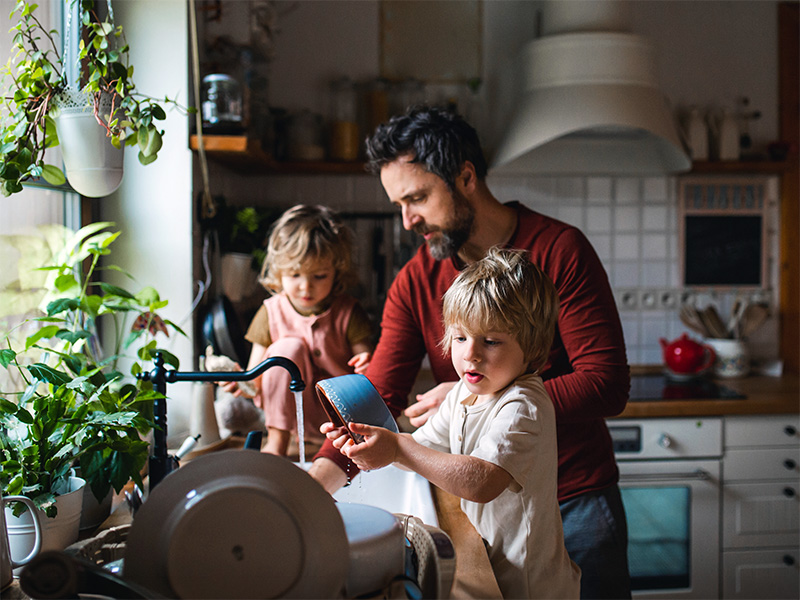Hombre y dos niños pequeños friegan los platos juntos junto a la ventana de la cocina.