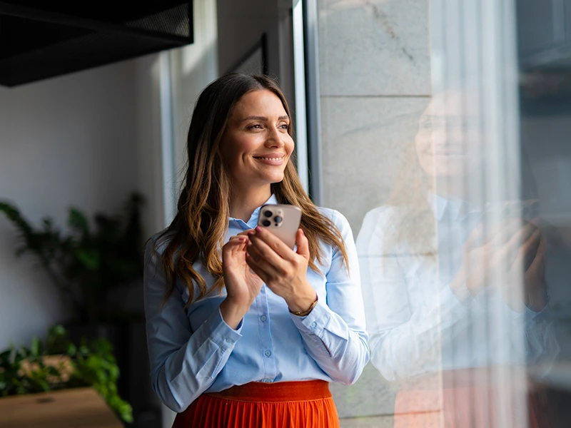 Mujer sonriente con un teléfono móvil en las manos, mirando por una ventana.
