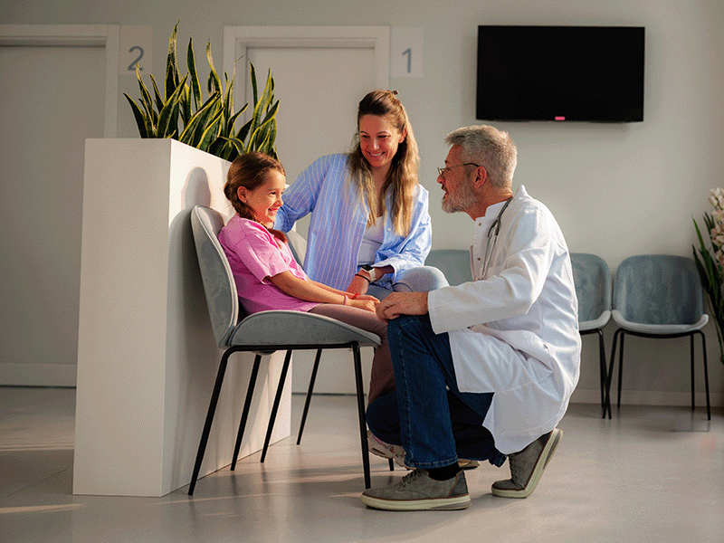 Profesional de la salud agachado conversa con una niña sonriente y una mujer en una sala de espera.